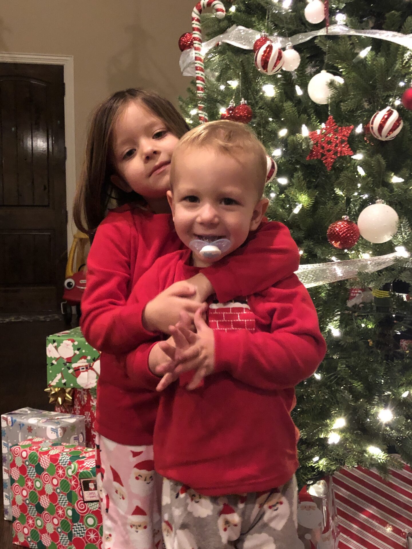 Two toddler kids in front of a Christmas tree. Older girl with arms wrapped around younger boy who has a pacifier hanging from his mouth.