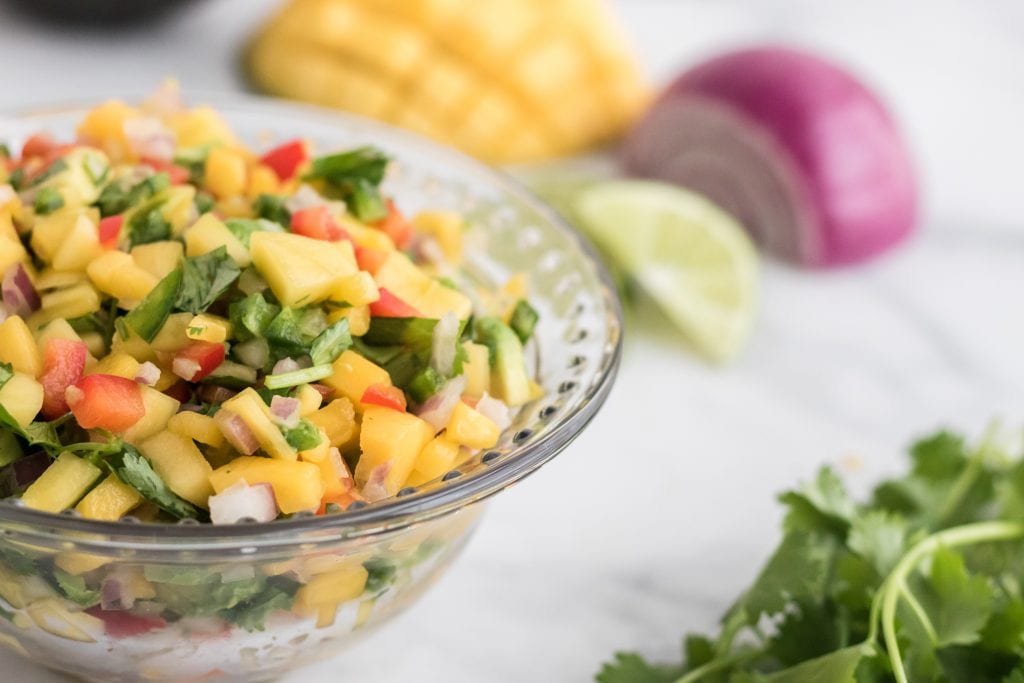 fresh mango salsa in bowl with onion, lime, mango, and cilantro in background