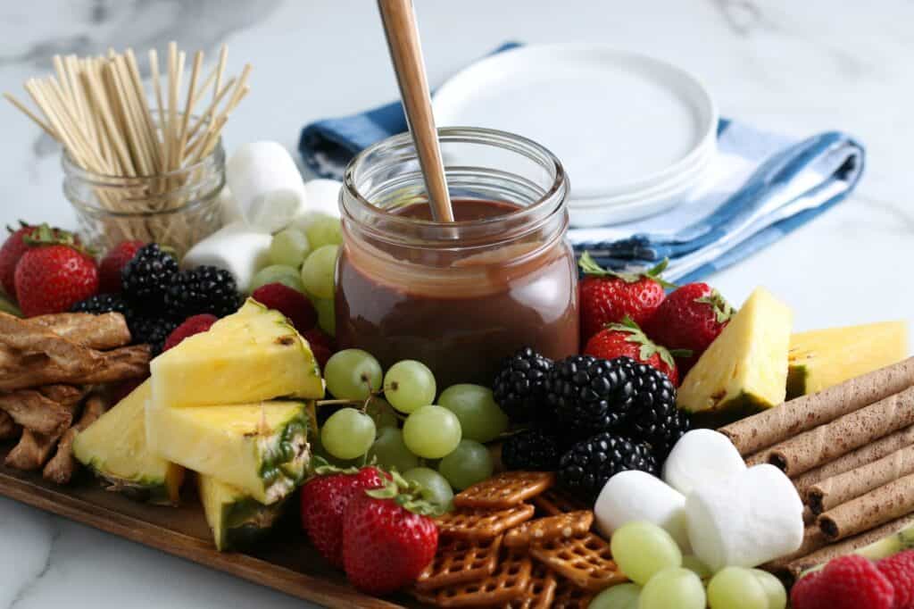 The chocolate sauce in a jar surrounded with fruit, pretzels, and marshmallows with a jar of toothpicks in the background.