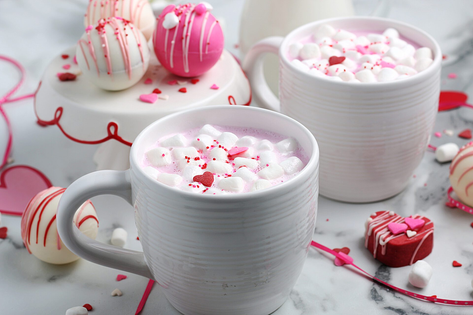pink hot chocolate with marshmallows in white mugs surrounded by hot chocolate bombs