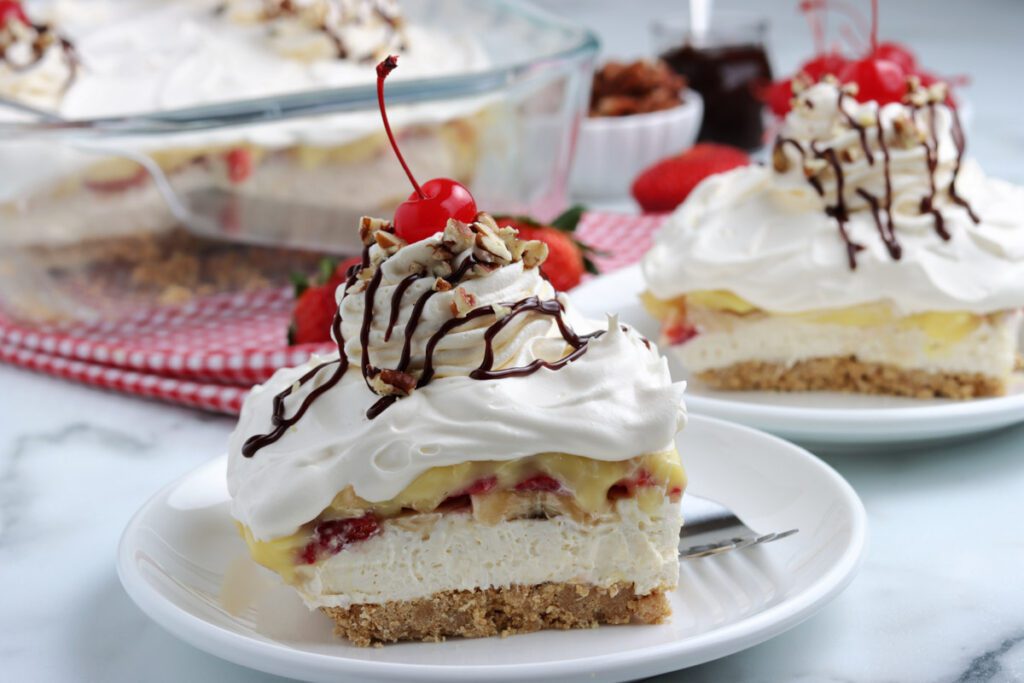 close up of banana split cake topped with whipped cream, chocolate, nuts, and cherry