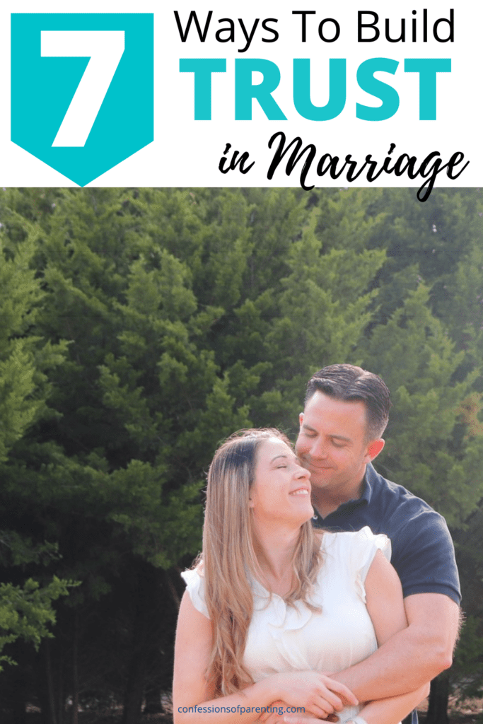 Man holding his wife from behind, smiling at her. Wife has eyes closed and smiling. The couple is in front of a pine tree background.