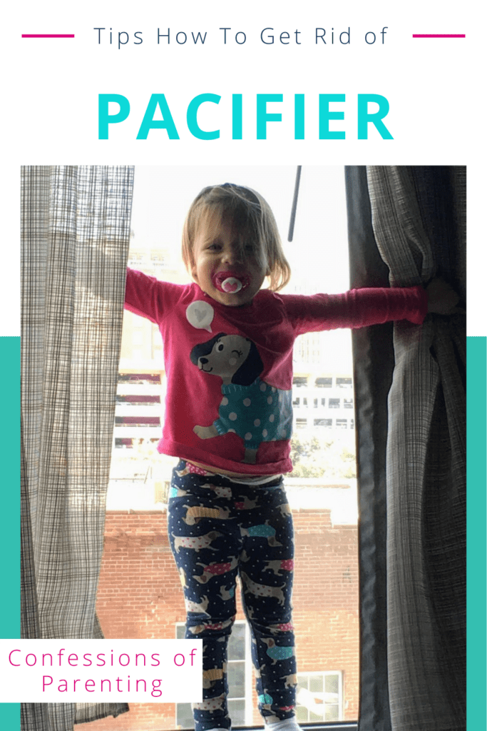 Toddler girl standing on a window sill with hands on curtains and pacifier in her mouth.