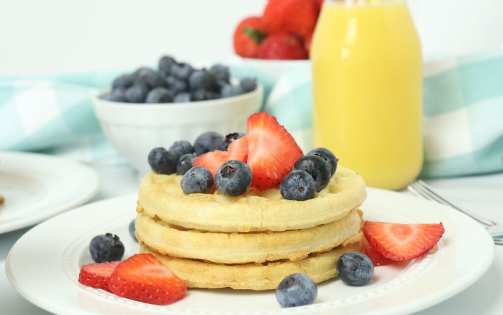 eggo waffles topped with berries on white plate with orange juice in glass behind it. with blue linen. 