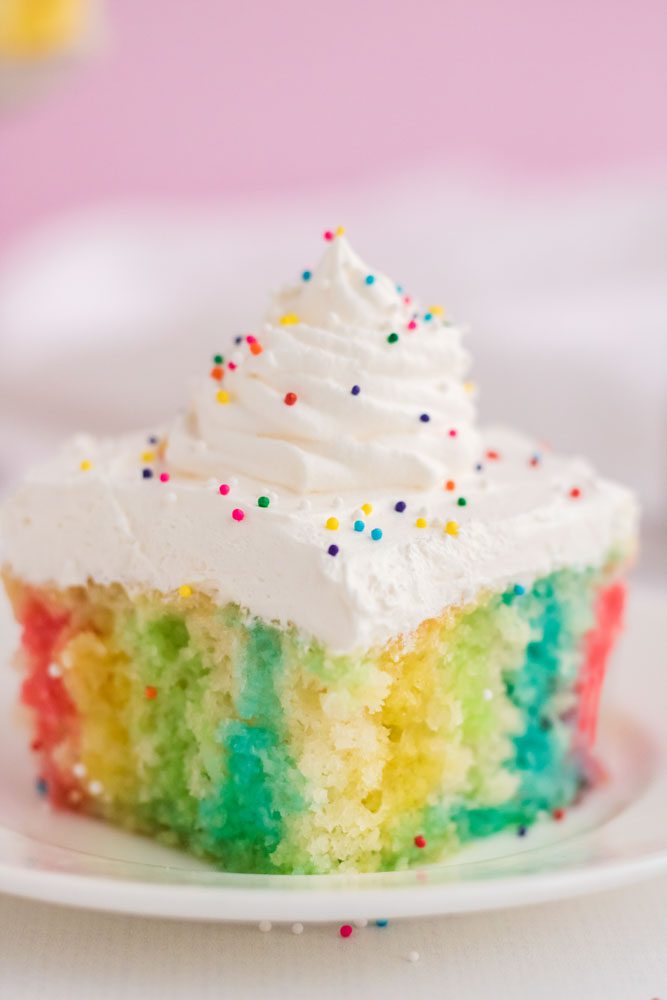 close up of rainbow poke cake on plate with sprinkles