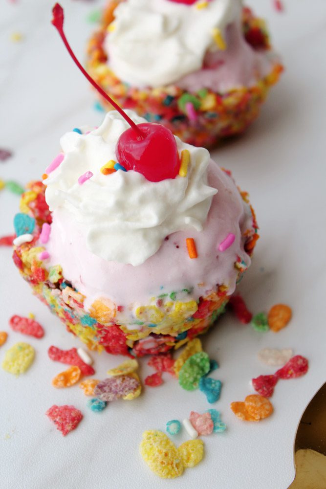 rainbow cereal ice cream cups with strawberry ice cream with whipped cream and cherry inside on white dish. 