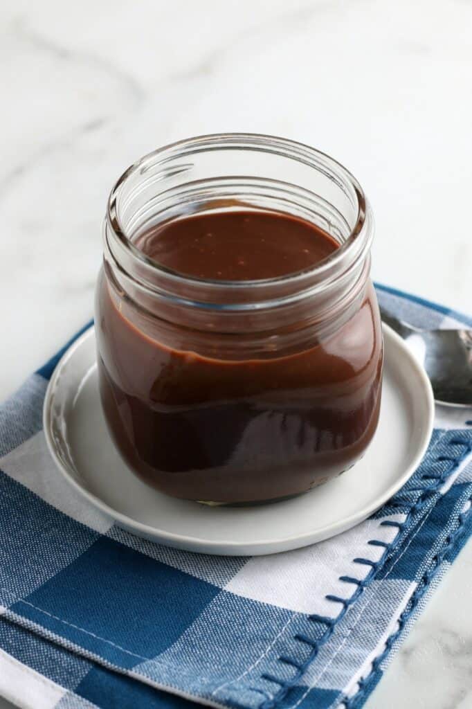 The chocolate sauce in a small jar with a spoon and napkin in the background. 