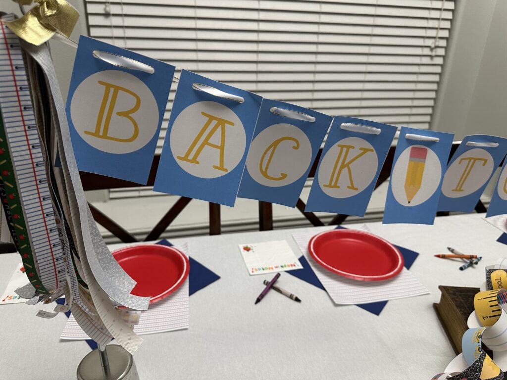 Close-up of the “BACK” portion of a back to school banner with bold yellow letters on blue cards, strung with white ribbon and surrounded by festive ribbons.