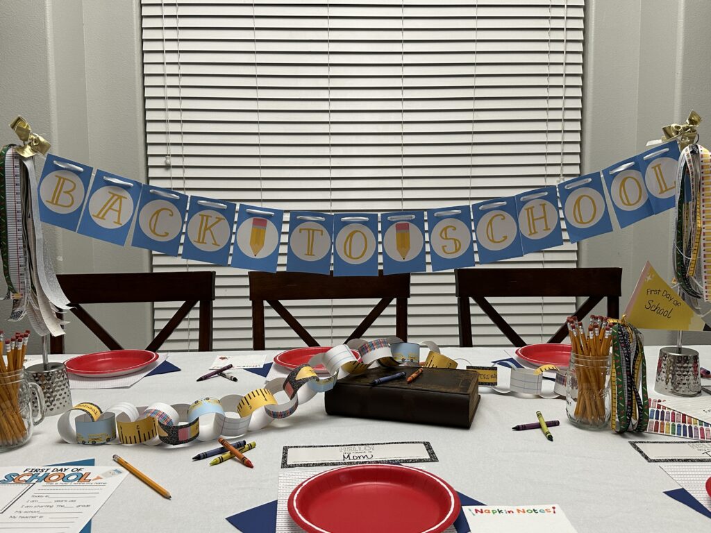 Full view of a decorated dining table with a “BACK TO SCHOOL” banner hanging above, complete with red plates, pencils, ribbon bundles, and a first day of school chain centerpiece.