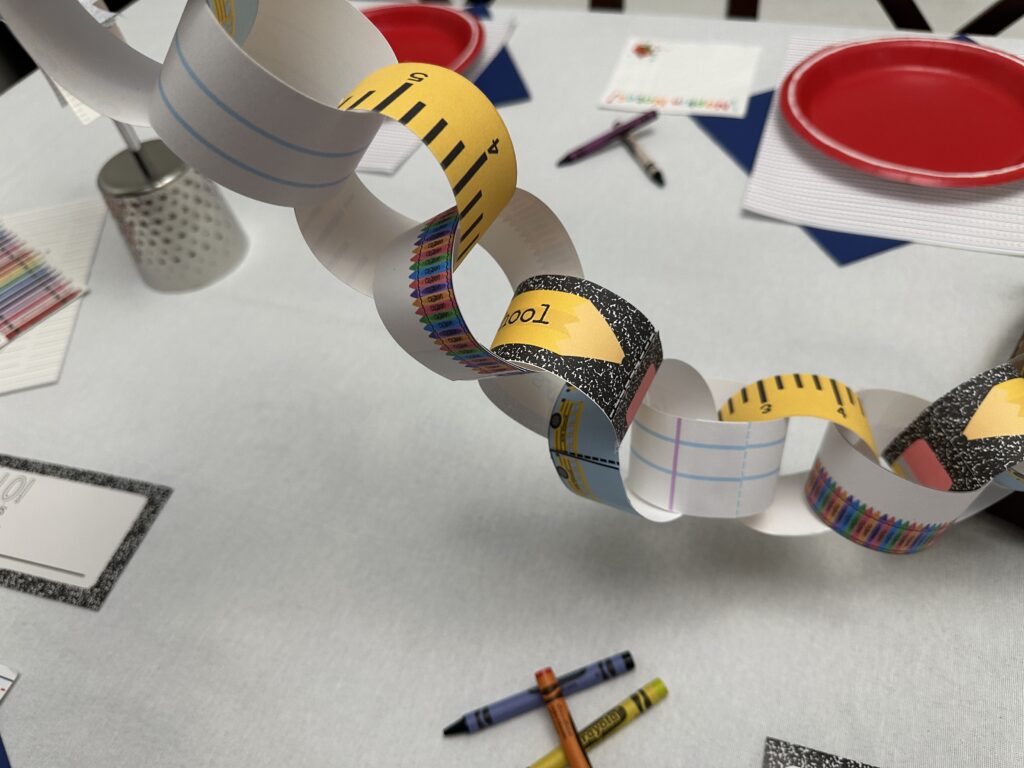 Back to school Countdown chain hanging from above with visible numbered ruler strips and composition notebook designs, surrounded by crayons and red plates.