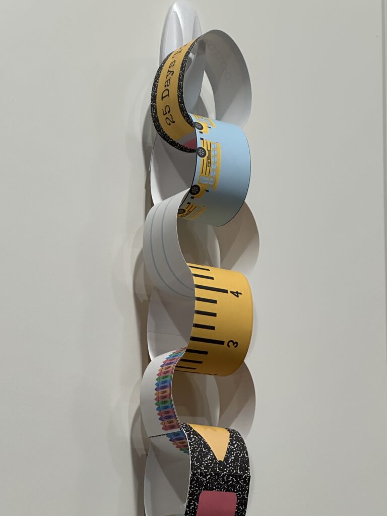 A close-up view of a colorful first day of school countdown chain hanging from a hook, featuring school-themed designs like buses, rulers, notebook paper, and crayons.