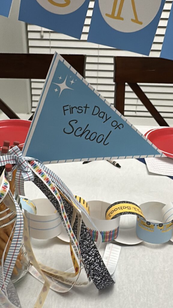Blue "First Day of School" flag centerpiece with decorative school-themed ribbons and a colorful paper countdown chain on a white table.