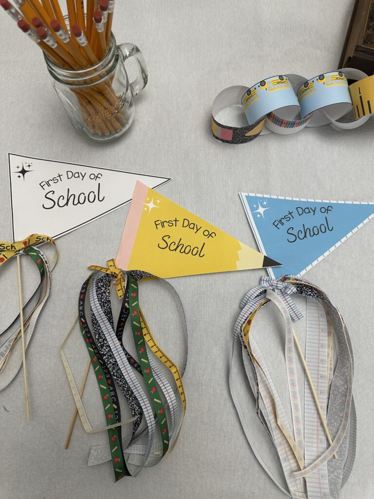 Overhead view of completed blue and yellow "First Day of School" flags with ribbon tails, laying on a white table next to a paper chain and mason jar of pencils.