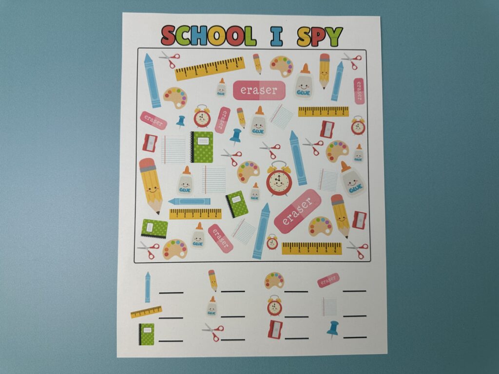 Flat lay image of a colorful "School I Spy" printable activity on a blue background. The page is filled with cute school-themed illustrations like crayons, pencils, rulers, glue, scissors, erasers, notebooks, and alarm clocks for kids to find and count.