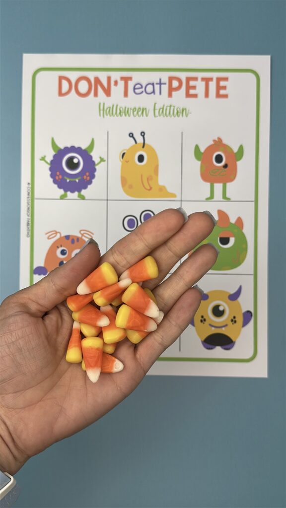 A hand holding several pieces of candy corn over a printable Halloween game board titled "Don't Eat Pete – Halloween Edition," featuring cute monster illustrations in a 3x3 grid.