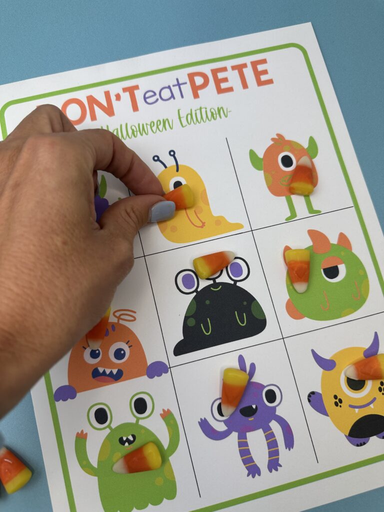 A hand picking up a piece of candy corn from a monster square on the "Don't Eat Pete – Halloween Edition" printable game board, with candy corn covering multiple monsters.