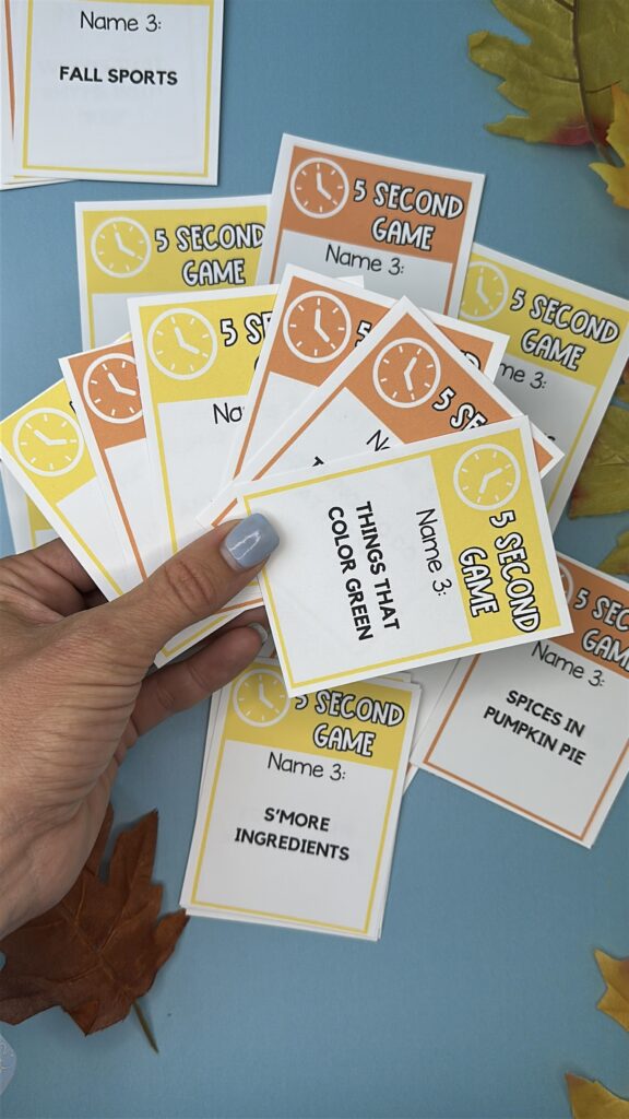 Hand holding colorful fall-themed 5 Second Game cards with prompts like “S’more ingredients” and “Things that color green.”