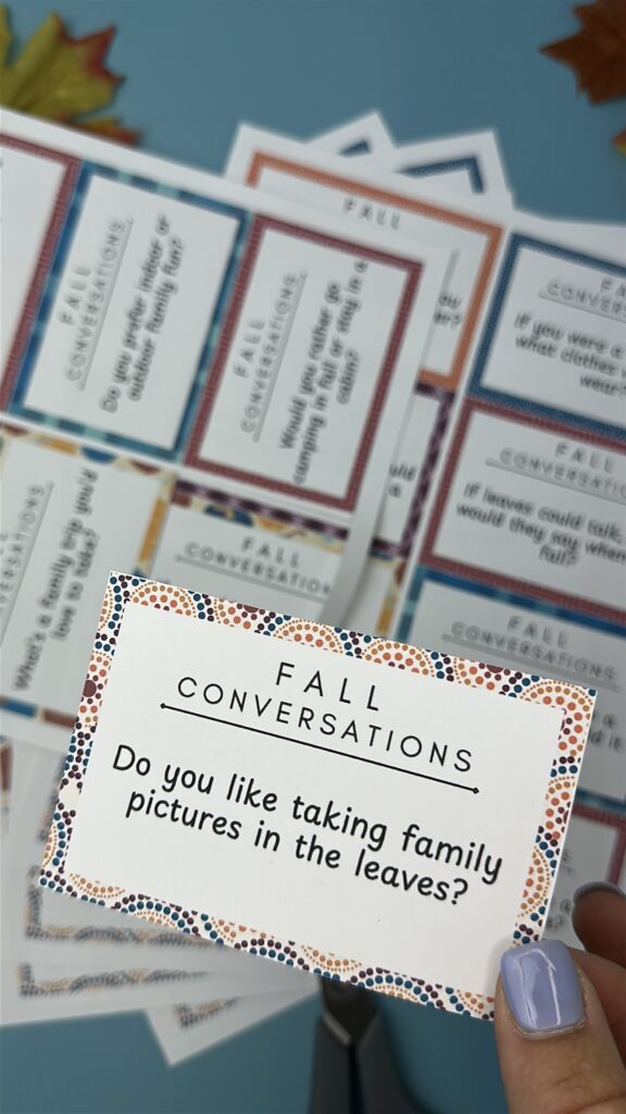 Close-up of a hand holding a fall conversation starter card that says, “Do you like taking family pictures in the leaves?” More printable cards with colorful borders are spread out in the background.