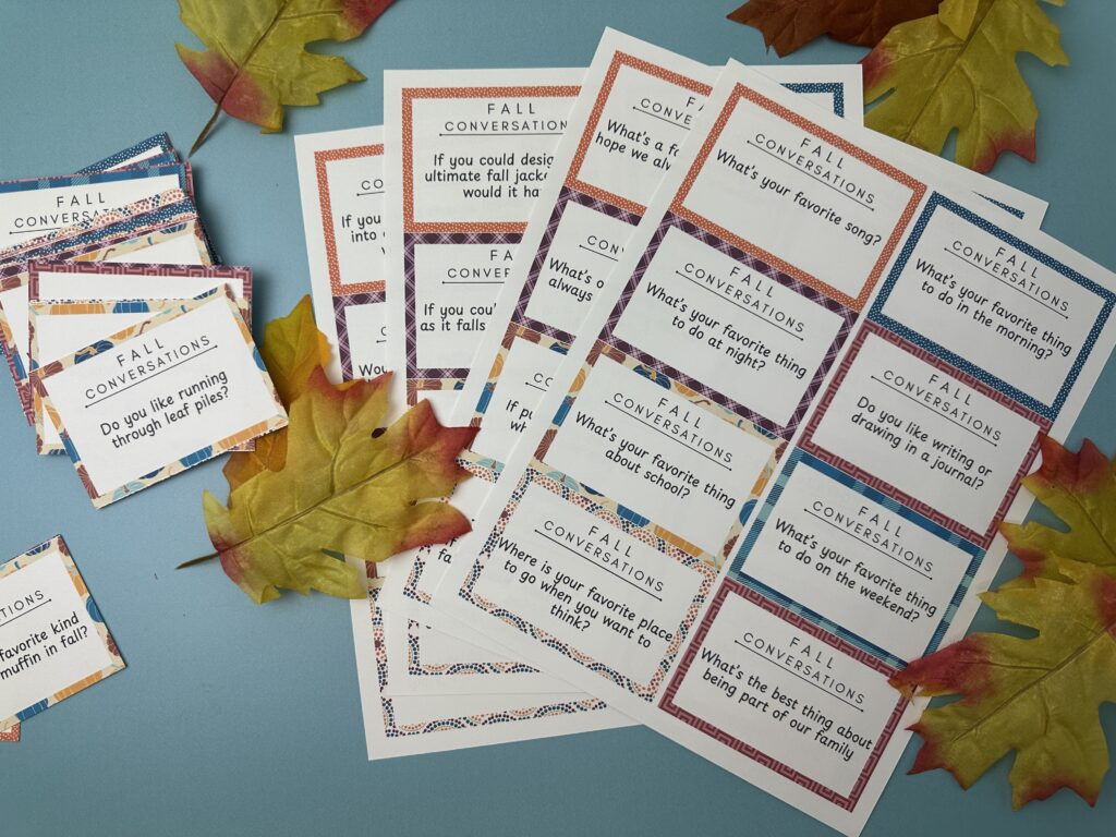 Sheets of printable fall conversation starter cards spread out on a blue background with decorative autumn leaves. Prompts include “Do you like running through leaf piles?” and “What’s your favorite thing about school?”