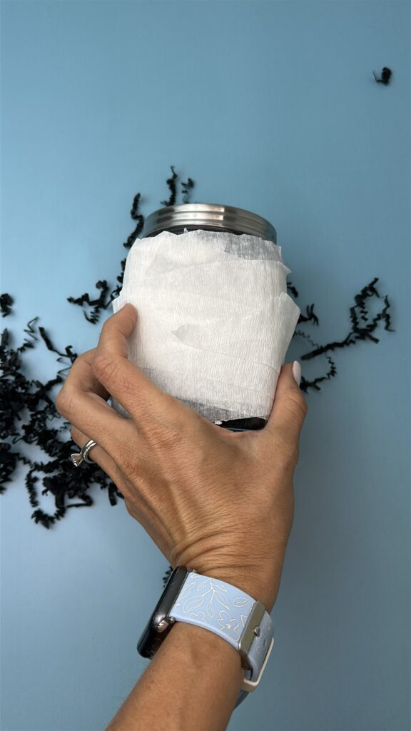 Hand wrapping white crepe paper around a candy jar to create a mummy gift jar.
