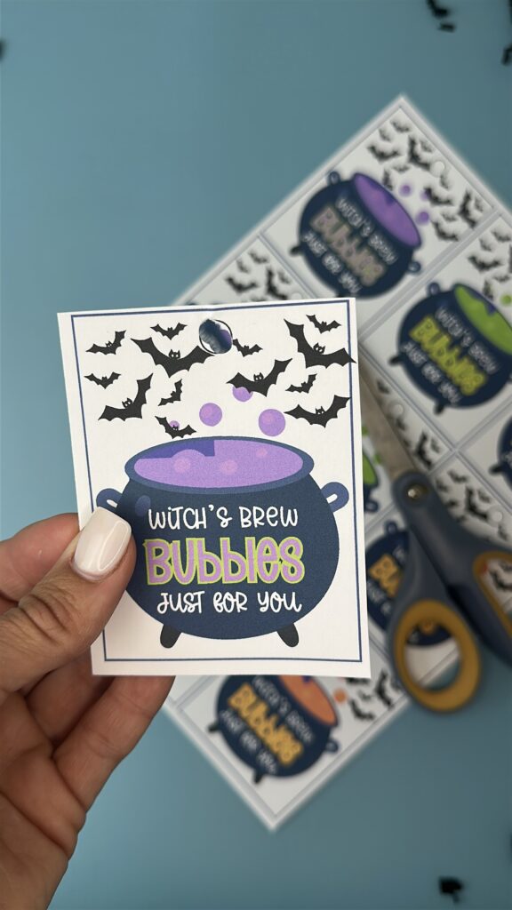 A hand holding a Halloween printable tag featuring a bubbling purple cauldron with the words "Witch’s Brew Bubbles Just for You," surrounded by flying bats. Other cut-out tags and scissors are visible in the background.