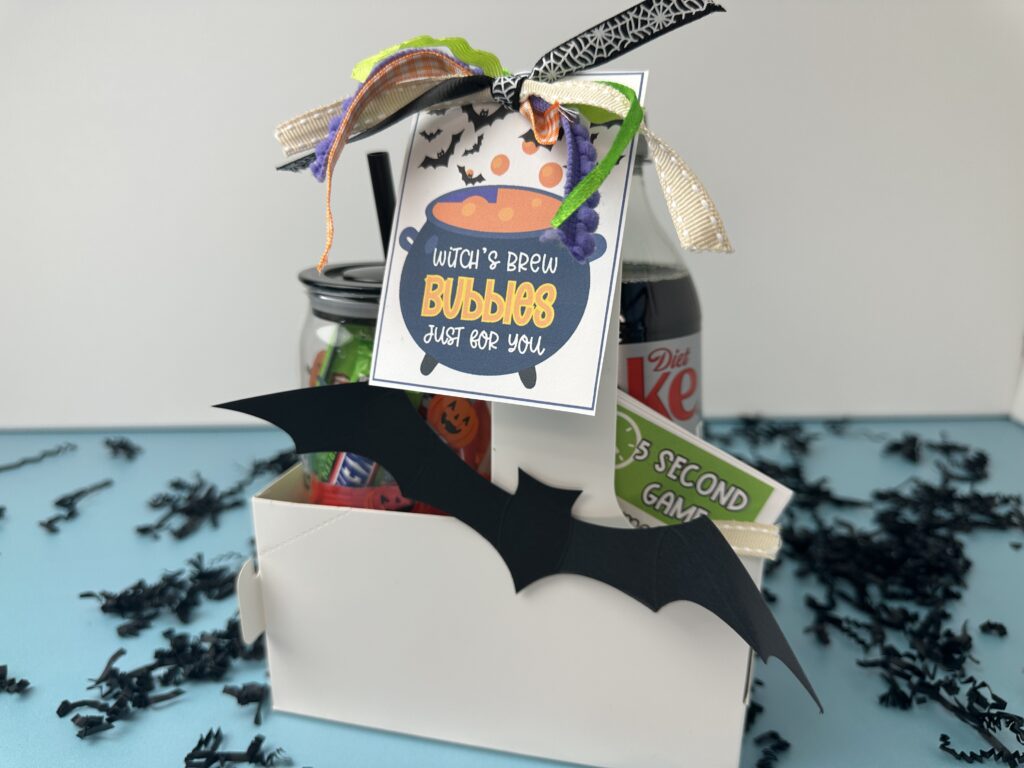A white Halloween gift box filled with soda and goodies, decorated with a black paper bat and tied with colorful ribbon. A tag on the front reads "Witch’s Brew Bubbles Just for You."