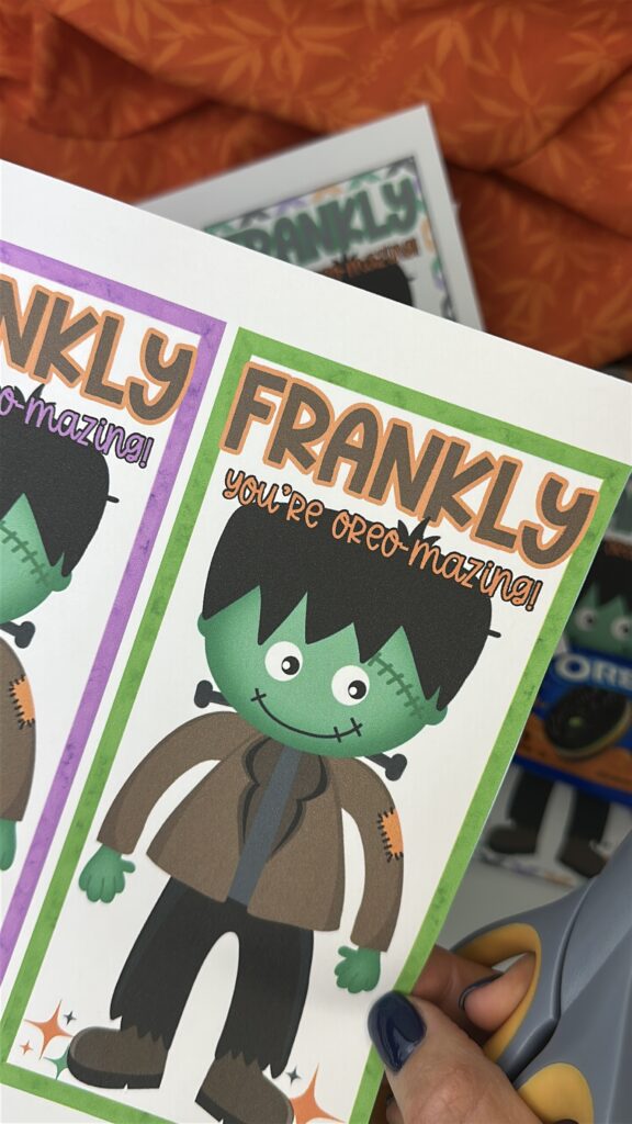 Close-up of printable Frankenstein treat tags reading “Frankly, You’re Oreo-Mazing!” held by hand with scissors nearby.