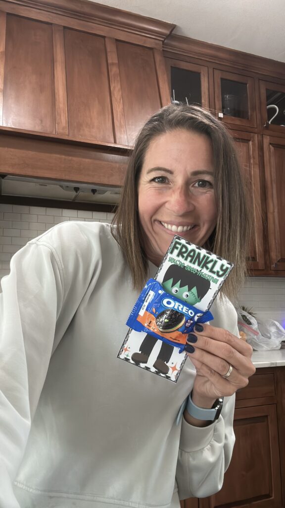 Smiling Michele Tripple holding Frankenstein Oreo treat card labeled “Frankly, You’re Oreo-Mazing!” in kitchen setting.