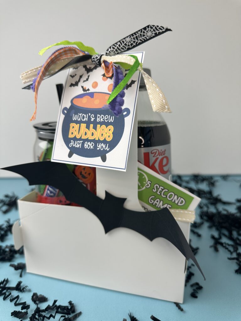 Close-up front view of the Halloween gift box with Diet Coke, Halloween treats, ribbon, and a bat decoration. A colorful tag with a bubbling orange cauldron reads "Witch’s Brew Bubbles Just for You."