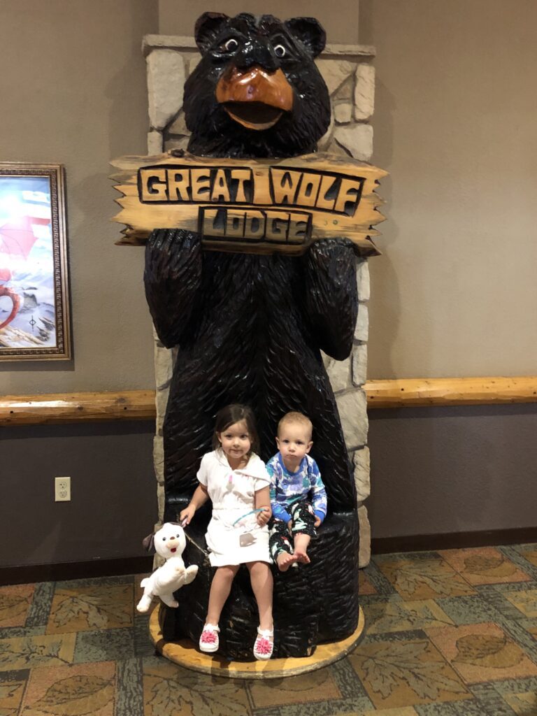 Two young children sitting by the Great Wolf Lodge bear statue holding a sign that says “Great Wolf Lodge,” smiling at the start of their family vacation.
