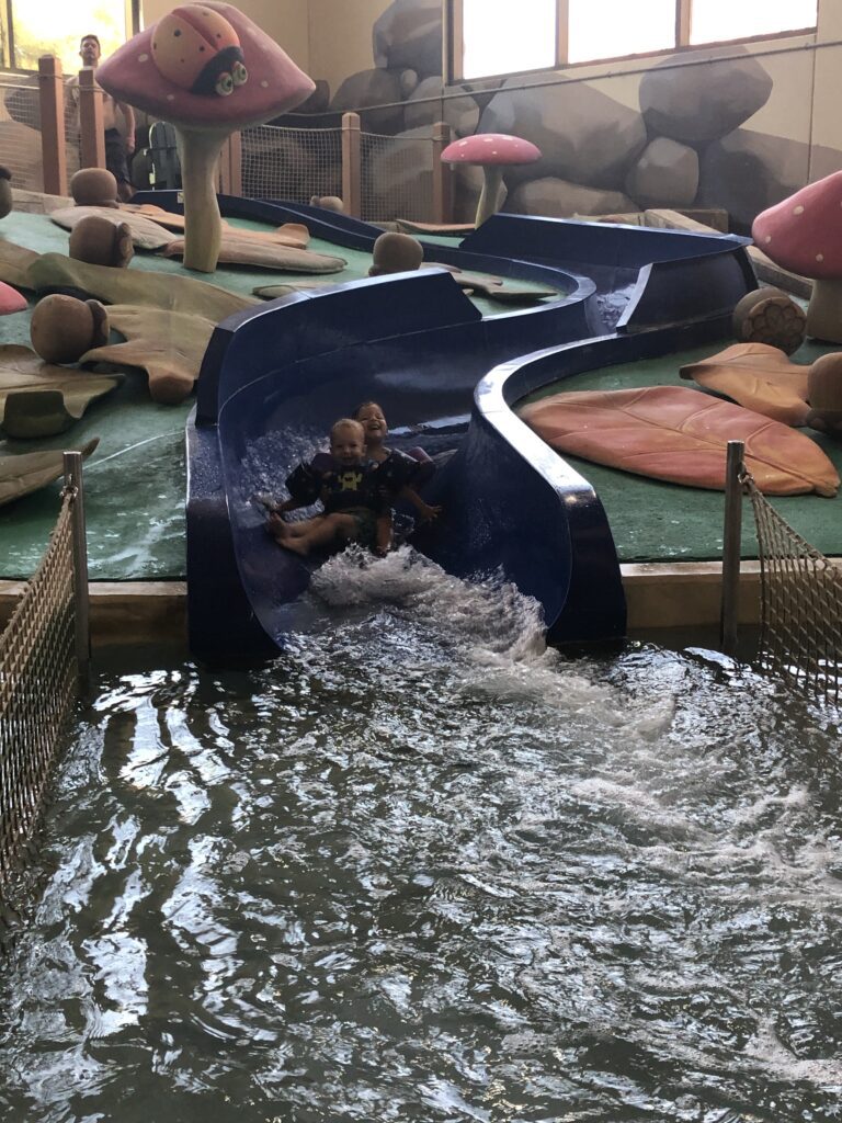 Young kids sliding down a small blue slide into the toddler pool area at Great Wolf Lodge indoor water park in Texas.
